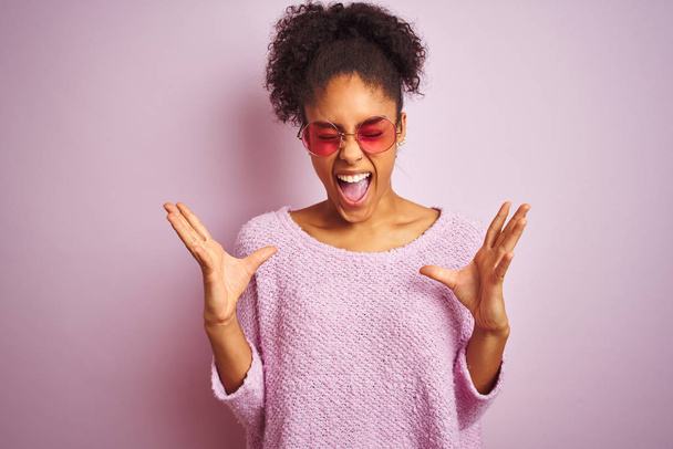 Mujer afroamericana con suéter de invierno y gafas de sol sobre fondo rosa aislado celebrando loco y loco por el éxito con los brazos levantados y los ojos cerrados gritando emocionados. Concepto ganador
 - Foto, imagen