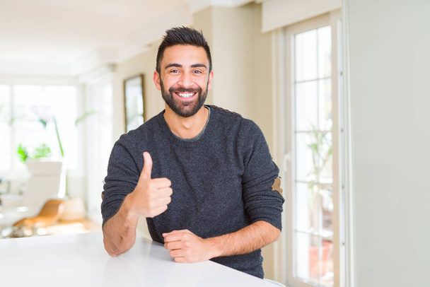 Hombre hispano guapo usando suéter casual en casa haciendo un gesto feliz con la mano. Aprobar la expresión mirando a la cámara con éxito
. - Foto, imagen
