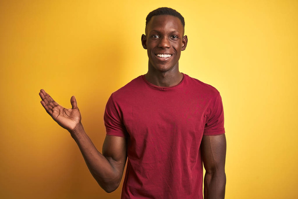 Joven hombre afroamericano vistiendo camiseta roja de pie sobre un fondo amarillo aislado sonriendo alegre presentando y señalando con la palma de la mano mirando a la cámara
. - Foto, Imagen
