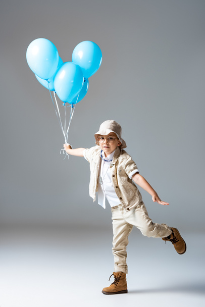 vista a figura intera del bambino esploratore in occhiali in piedi su una gamba e tenendo palloncini blu su grigio
 - Foto, immagini