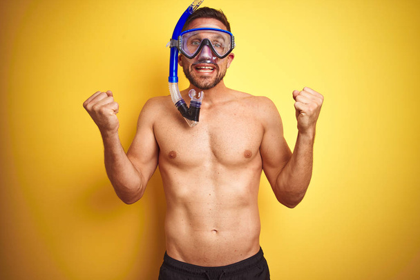 Young handsome shirtless man wearing diving goggles over isolated yellow background screaming proud and celebrating victory and success very excited, cheering emotion - 写真・画像