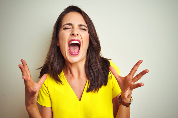 Young beautiful woman wearing yellow t-shirt standing over white isolated background crazy and mad shouting and yelling with aggressive expression and arms raised. Frustration concept. - Fotografie, Obrázek
