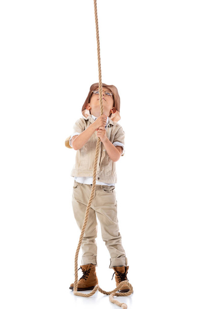full length view of explorer kid in hat and glasses holding rope and looking up on white - Фото, зображення