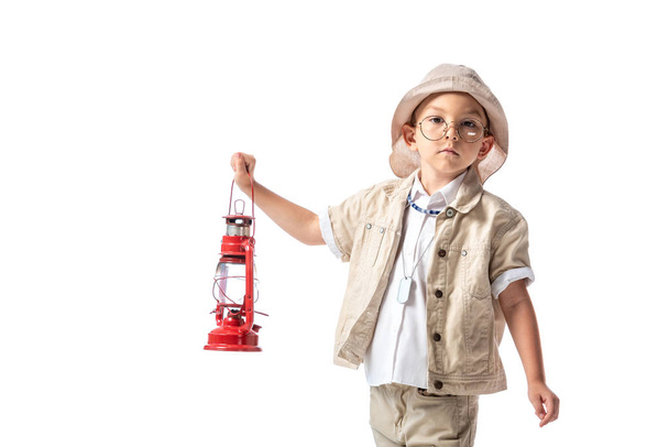 pensive explorer boy in glasses and hat holding red lantern and looking at camera isolated on white - 写真・画像