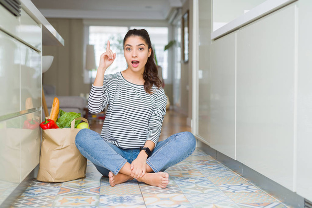 Молодая женщина, сидящая на полу кухни с бумажным пакетом, полным свежих продуктов, указывая пальцем вверх с успешной идеей. Взволнован и счастлив. Номер один
. - Фото, изображение