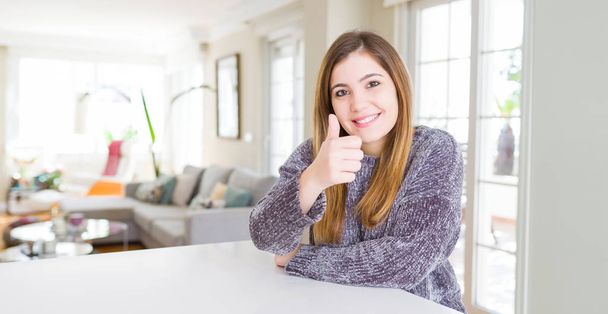 Beautiful young woman at home doing happy thumbs up gesture with hand. Approving expression looking at the camera showing success. - Fotografie, Obrázek
