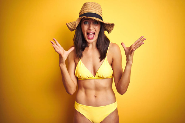 schöne Frau in gelbem Bikini und Sommermütze über isoliertem gelben Hintergrund feiert verrückt und erstaunt für den Erfolg mit erhobenen Armen und offenen Augen schreit aufgeregt. Siegerkonzept - Foto, Bild