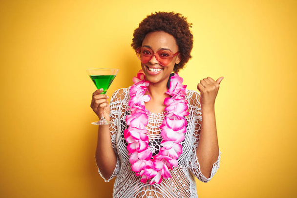 Joven mujer afroamericana con pelo afro vistiendo lei hawaiano flor y bebiendo un cóctel señalando y mostrando con el pulgar hacia el lado con la cara feliz sonriendo
 - Foto, imagen