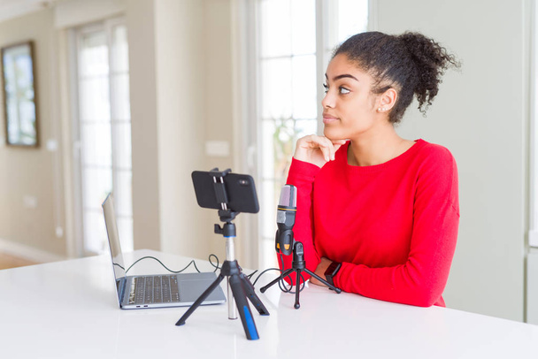 Jonge Afro-Amerikaanse vrouw doet video-oproep met behulp van smartphone camera en microfoon met de hand op kin denken over vraag, pensieve uitdrukking. Glimlachend met doordachte gezicht. Doubt concept. - Foto, afbeelding