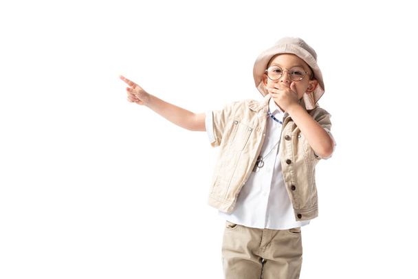 σοκαρισμένος εξερευνητής αγόρι σε καπέλο και γυαλιά καλύπτοντας το στόμα με το χέρι και δείχνοντας με το δάχτυλο απομονώνεται σε λευκό - Φωτογραφία, εικόνα