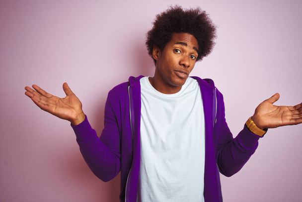 Mladý africkej Američan v purpurovém triku, který stál nad izolovaným růžovým pozadím, beztvarý a zmatený, se zdviženýma rukama. Koncepce pochybností. - Fotografie, Obrázek