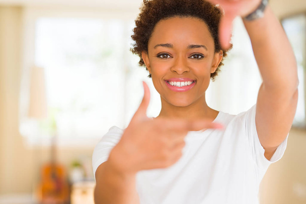Jeune belle femme afro-américaine portant un t-shirt blanc décontracté souriant faisant cadre avec les mains et les doigts avec le visage heureux. Concept de créativité et de photographie
. - Photo, image