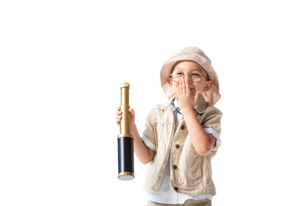 smiling explorer boy in glasses and hat holding spyglass and covering mouth with hand isolated on white - Fotoğraf, Görsel