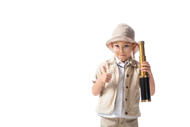 front view of smiling explorer boy in glasses and hat holding spyglass and showing thumb up isolated on white - Photo, Image
