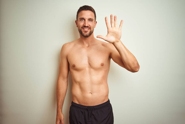 Homem sem camisa bonito jovem sobre fundo isolado mostrando e apontando para cima com os dedos número cinco, sorrindo confiante e feliz
. - Foto, Imagem