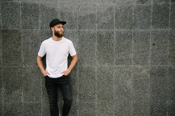 Hipster handsome male model with beard wearing white blank t-shirt and a baseball cap with space for your logo or design in casual urban style - Fotoğraf, Görsel
