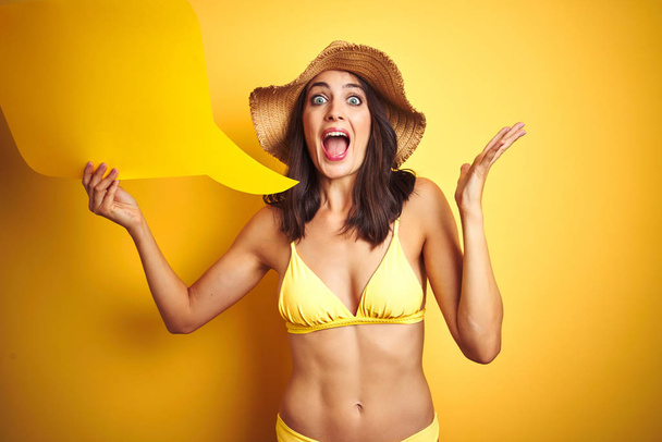 Mooie vrouw dragen gele bikini en houden praten ballon over geïsoleerde gele achtergrond erg blij en opgewonden, winnaar uitdrukking vieren overwinning schreeuwen met grote glimlach en verhoogde handen - Foto, afbeelding
