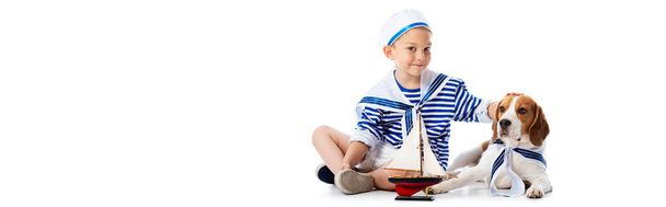 白にビーグル犬をなでるおもちゃの船でセーラー服を着た笑顔の少年のパノラマショット - 写真・画像