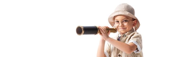 panoramic shot of smiling explorer boy in hat and glasses holding spyglass isolated on white - Фото, изображение