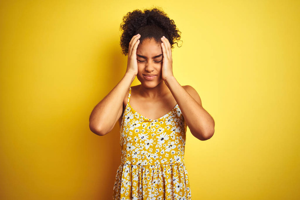 Africká Američanka nosí neformální květinové šaty stojící nad izolovanými žlutými pozadí trpícími bolestmi bolestí hlavy a vystresovanými, protože bolest a migréna. Ruce na hlavu. - Fotografie, Obrázek