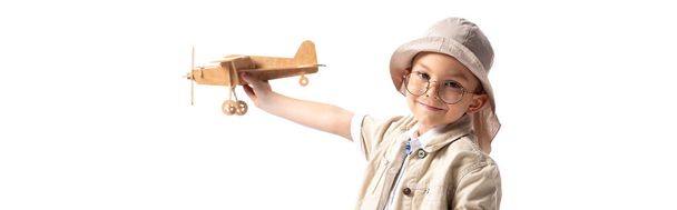 panoramic shot of smiling explorer child in glasses and hat holding wooden toy plane isolated on white - Photo, Image
