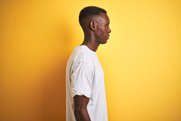 Young african american man wearing white t-shirt standing over isolated yellow background looking to side, relax profile pose with natural face with confident smile. - Foto, Imagem