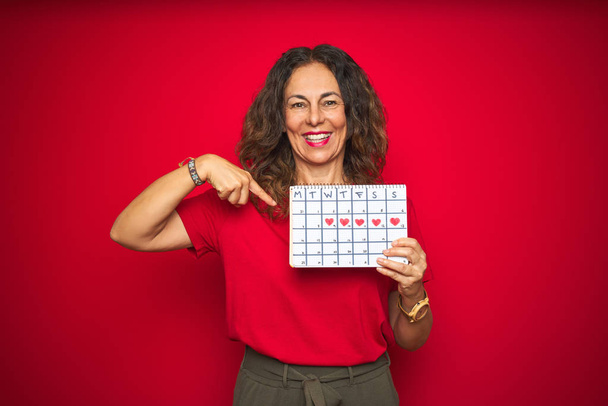 Старша жінка середнього віку, що тримає календар менструацій над червоним ізольованим тлом з несподіваним обличчям, спрямованим пальцем до себе
 - Фото, зображення