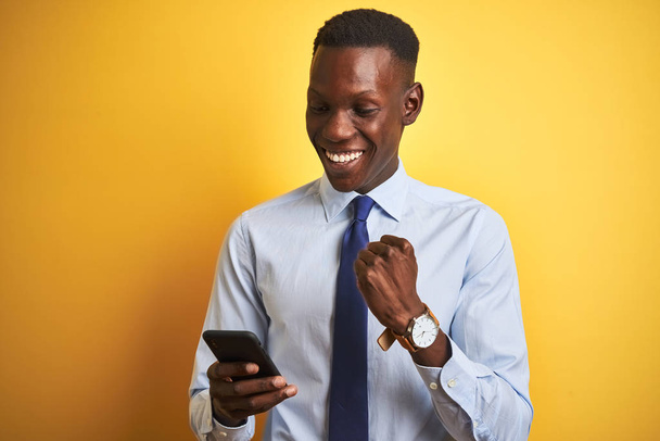 Afrikanischer amerikanischer Geschäftsmann mit Smartphone vor isoliertem gelben Hintergrund, der stolz schreit und Sieg und Erfolg feiert, sehr aufgeregt, jubelnde Emotion - Foto, Bild