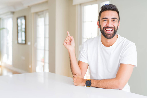 Handsome hispanic man casual white t-shirt at home with a big smile on face, pointing with hand and finger to the side looking at the camera. - Photo, Image