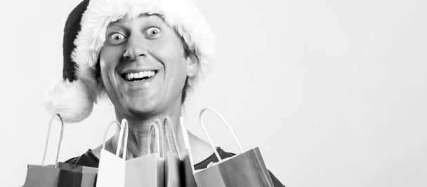 Aufgeregter junger Mann mit Weihnachtsmütze und Einkaufstüten. Weihnachten, Verkauf, Rabatt und Feiertage Konzept. weihnachtsverrückter Shopper mit Geschenken, auf weißem Hintergrund, Kopierraum. - Foto, Bild