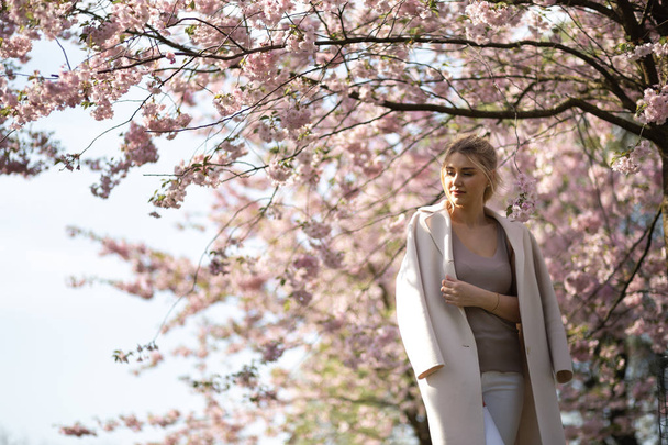 Jovem loira bonita no parque Sakura Cherry Blossom na primavera desfrutando da natureza e tempo livre durante o seu tempo livre de turista viajante - vestindo calças brancas e camiseta com uma jaqueta bege
 - Foto, Imagem