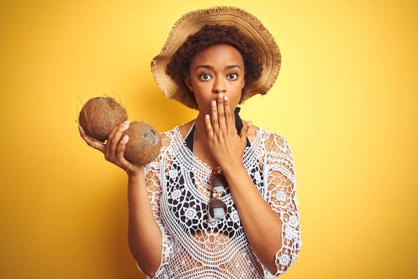 Joven mujer afroamericana con cabello afro sosteniendo coco sobre fondo amarillo aislado cubrir la boca con la mano conmocionada con vergüenza por error, expresión de miedo, asustado en silencio, concepto secreto
 - Foto, Imagen