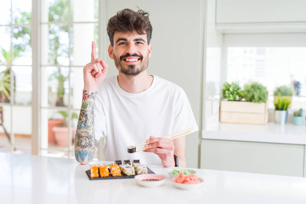 Молодой человек, поедающий азиатские суши из доставки на дом, удивлен идеей или вопросом, указывая пальцем со счастливым лицом, номер один
 - Фото, изображение