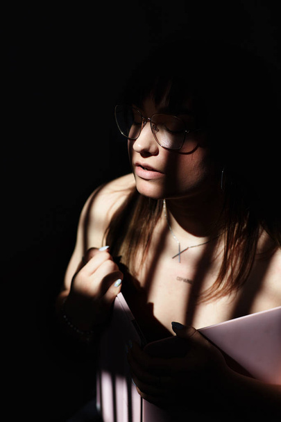 vertikale Aufnahme einer modischen jungen Frau mit dunklen, glatten, glänzenden Haaren, geschlossenen Augen, meditierend mit großer Brille, Armbänder und Armbanduhren tragend, auf dunklem Hintergrund posierend, Sonnenstrahlen im Gesicht einfangend - Foto, Bild