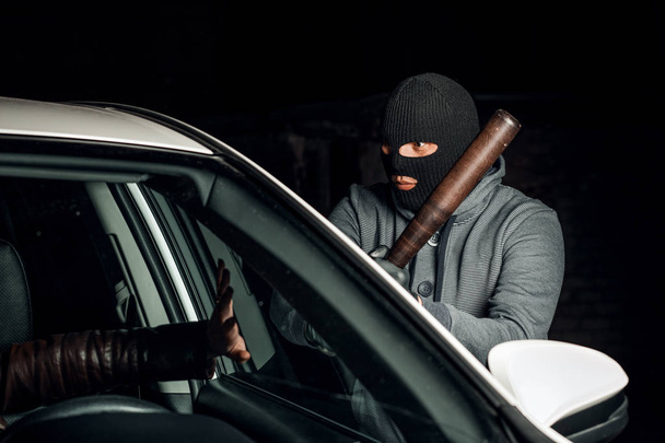robber in a Balaclava tries to break the car glass with a baseba - Photo, Image