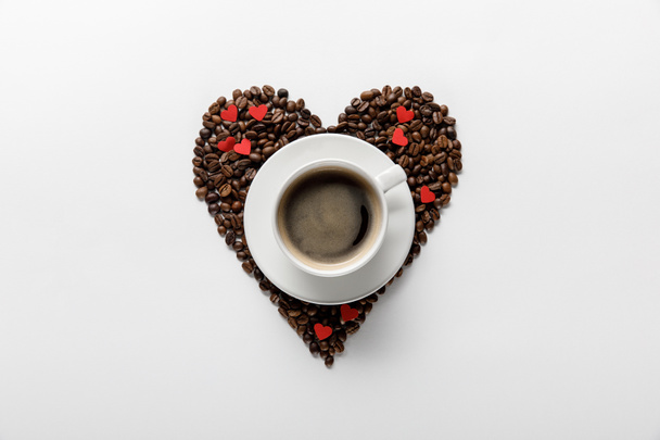 top view of coffee in cup on saucer with heart made of coffee grains on white background - Photo, image