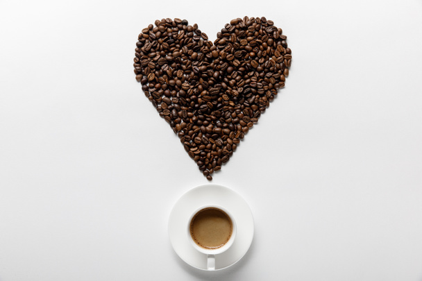 白い背景にコーヒーの穀物で作られた心を持つ受け皿のカップでおいしいコーヒーのトップビュー - 写真・画像
