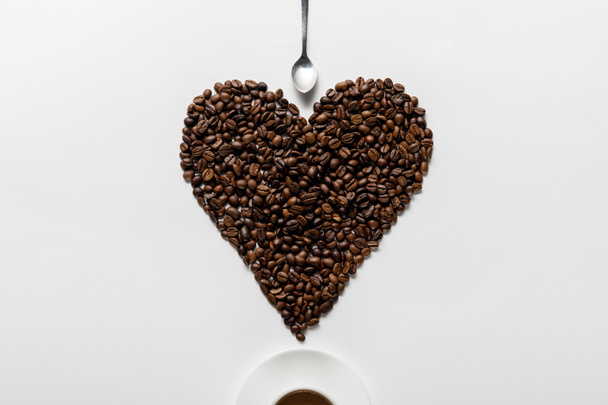 top view of coffee in cup on saucer with heart made of coffee grains near spoon on white background - Photo, Image