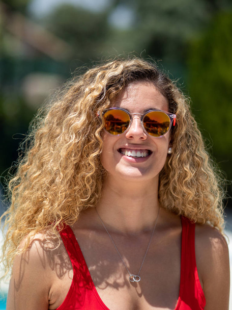 Chica rubia con pelo rizado y traje de baño rojo vista frontal con gafas de sol
. - Foto, Imagen