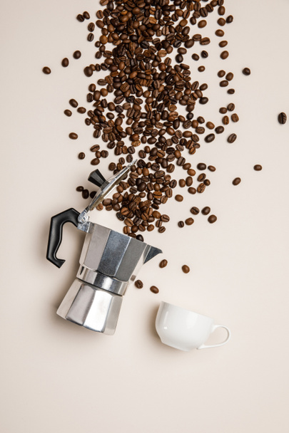 Top zicht op aluminium koffiepot, beker en verspreide verse koffiebonen op beige achtergrond - Foto, afbeelding