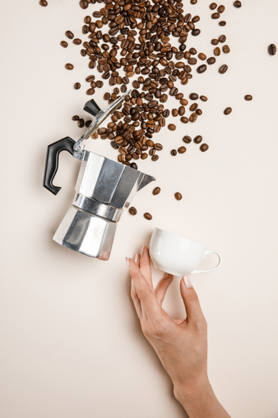partial view of woman holding cup near aluminium coffee pot and scattered fresh coffee beans on beige background - Foto, Imagem