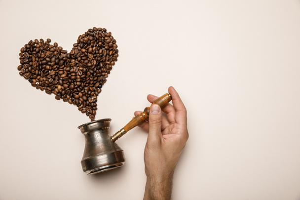 cropped view of man holding cezve near heart made of coffee grains on beige background - Photo, image