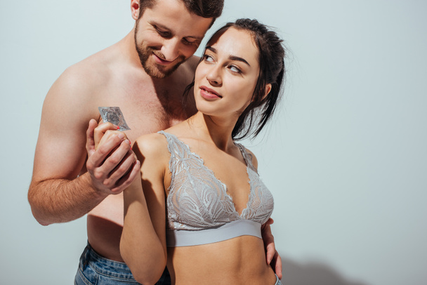 sexy pareja sonriendo y abrazando mientras chica sosteniendo condón y mirando al hombre
 - Foto, Imagen