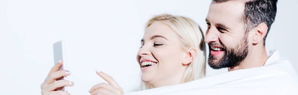 panoramic shot of man and beautiful girl covered in blanket using smartphone on white - Фото, зображення