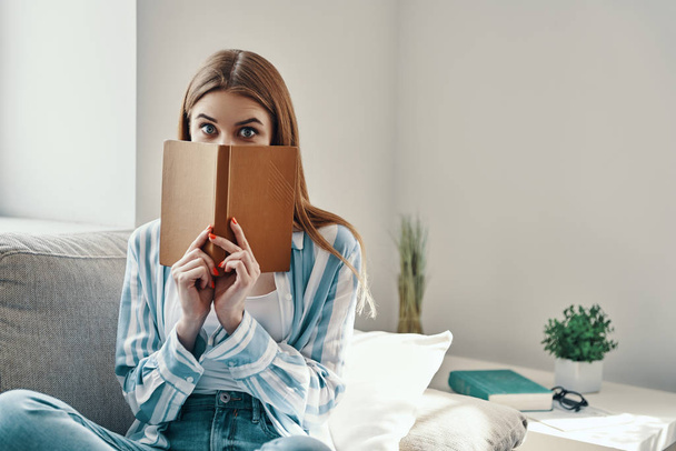 Attractive young woman looking at camera and covering face with a book while spending time at home - Valokuva, kuva