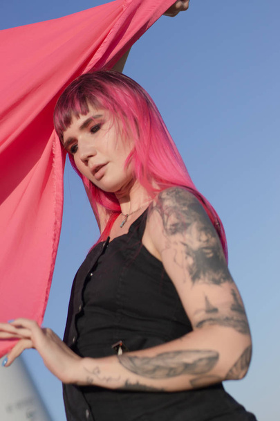Pink-haired young woman with pieces of pink cloth dance near the field with wind generators . Windmill on sunset. Fashion portrait - Фото, зображення