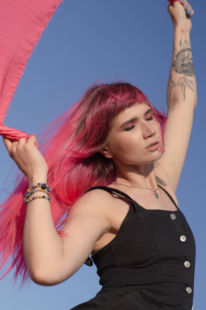 ピンクの髪の若い女性は、風の発電機でフィールドの近くでピンクの布の作品を踊ります.夕暮れ時の風車。ファッションの肖像画 - 写真・画像