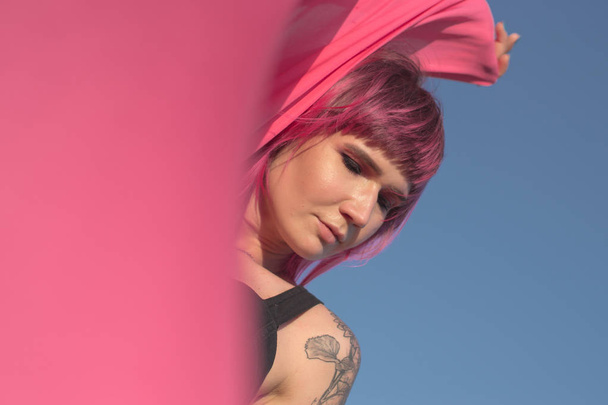 Pink-haired young woman with pieces of pink cloth dance near the field with wind generators . Windmill on sunset. Fashion portrait - Photo, Image