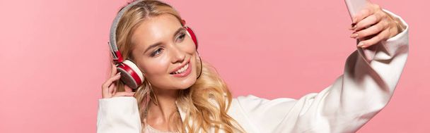 panoramic shot of beautiful blonde woman with headphones taking selfie on smartphone isolated on pink - Foto, imagen