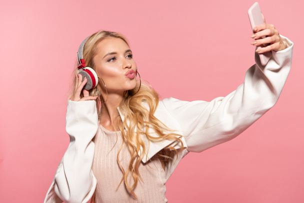schöne blonde Frau mit Kopfhörern macht Selfie auf dem Smartphone mit Entengesicht isoliert auf rosa - Foto, Bild